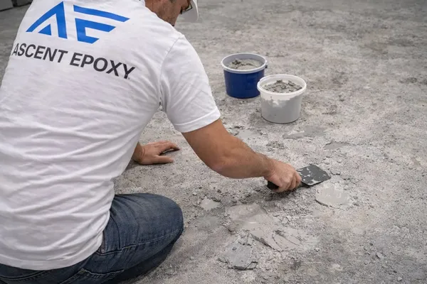 Crew member kneeling to spread patching compound into pitted concrete, two buckets of mix nearby